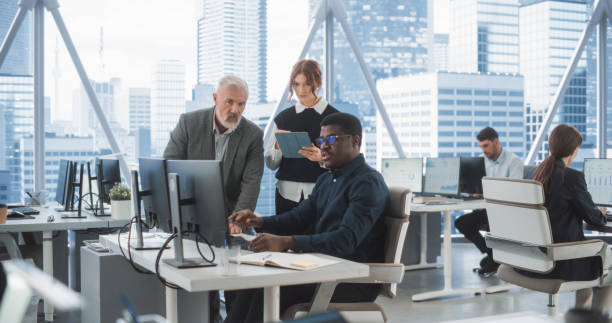 istock Diverse Colleagues Talking In Front Of Desktop Computer In Corporate Office With Megapolis Window View. Black Male Developer Showing New Code To Caucasian CEO And His Female Assistant With Tablet. 2075454559