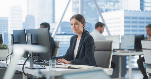 istock Focused Female Operations Specialist Typing On Desktop Computer In Diverse Corporate Office With Megapolis Window View. Professional Caucasian Woman Writing Emails to Clients, Scheduling Meetings. 2075454619