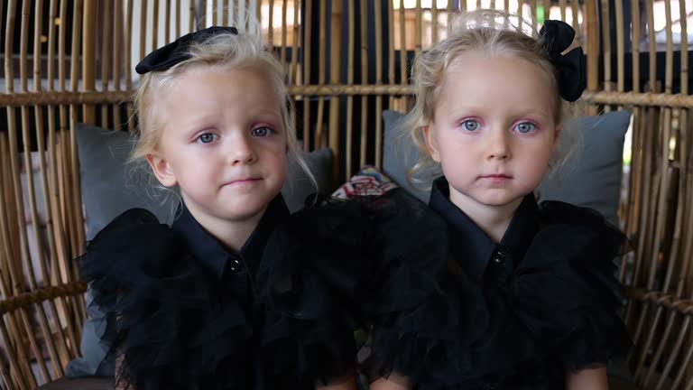 Two cute twin girls sitting in cafe in Wednesday suit with black bows on heads. Close-up of preschool kids came to cafe for Halloween party in Wednesday suits. Concept have fun and wear Wednesday suit