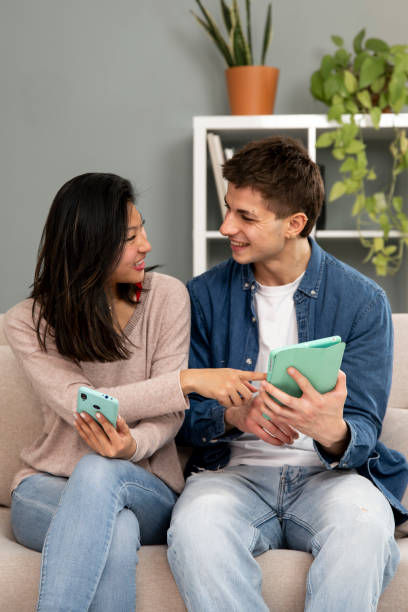 istock Diverse young couple smiling and having fun using online device. Smiling woman pointing tablet and man sitting on a couch in their living room at home. 2075456827