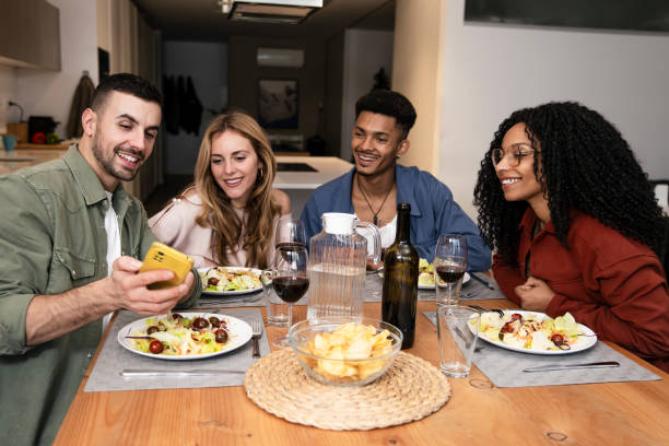 istock Smiling people sharing smartphone while eating on a modern apartment. Diverse group of happy friends having a video call diner at home. 2075460471