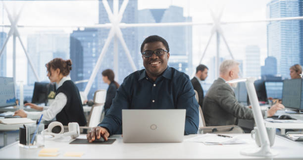 istock Succesful Black Man Typing On Laptop Computer In Bright Scyscraper Office With Diverse Employees In Downtown. Cheerful Male Programmer Looking At Camera And Smiling In Fintech Startup Workspace. 2075467289