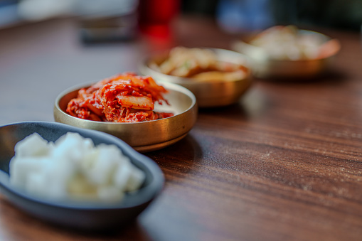 The vibrant colors and intricate textures of traditional Korean side dishes, prominently featuring kimchi, are showcased in this culinary photograph. The shallow depth of field accentuates the rich, spicy red hues of the fermented cabbage, evoking the authentic flavors of Korea, while blurred banchan in the background invites curiosity about the variety and complexity of Korean gastronomy.