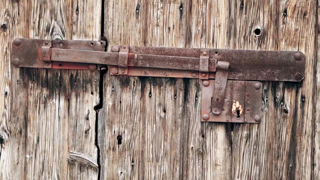 An old wooden door with a hinged vintage iron bolt. Rusty massive iron lock on an abandoned wooden gate. Sun-faded, aged and bark beetle-eaten wooden doors. Rovinj, Croatia - February 29, 2024