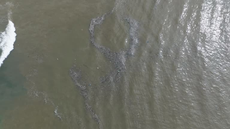 Slow motion high altitude top down drone shot of polluted ocean filled with floating trash and debris and muddy sewage runoff with green tropical forest and beach in  Bali Indonesia