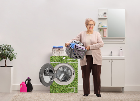 Full length portrait of a mature woman holding a laundry basket with clothes inside a bathroom with eco friendly washing machine