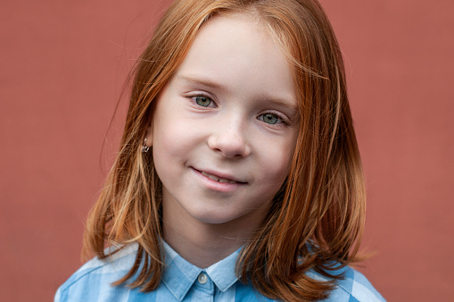 Portrait of a red-haired child with green eyes 8 years old in a blue shirt on a teracot background. The hair of the child is up to the shoulders. There is a soft smile on his face.