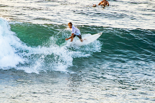 Bali, Uluwatu - Surfer Riding a Green Wave: A surfer on a white surfboard skillfully rides a mid-breaking green wave, while another person observes from the background. The ocean foam splashes around the surfer, capturing the dynamic motion of the ride.