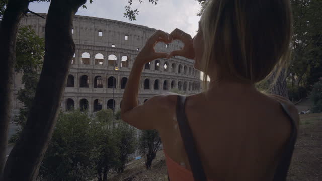 Young woman at the Colosseum in Rome making a heart shape finger frame with hands loving travel in Italy