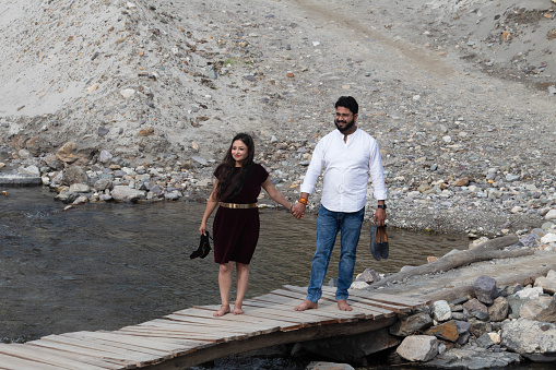 Couple strolls hand in hand on a footbridge over the Ganges River in Rishikesh, carrying their footwear as they gracefully traverse the bridge together, Uttarakhand
