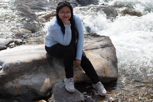 Joyous woman sits, relaxed with her hand on her chin, on an oversized rock by the gushing waters of the Ganges River in Rishikesh, smiling and looking directly at the camera, Uttarakhand