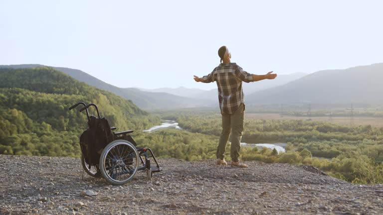 Portrait of incapacitated man which getting up from wheelchair on the mount and stretching arms and enjoying perfect feelings of freedom under sunlight