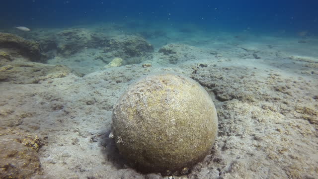 Close-up of an old stone cannonball lying on rocky seabed, on blue water background