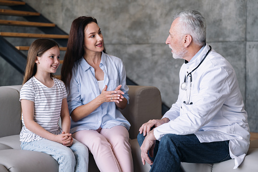 Family doctor talking to smiling mother and happy healthy child during home visit. Woman having conversation with friendly male pediatrician. Mum and daughter sitting on sofa and talking about health