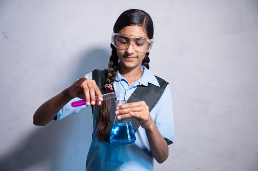 Happy Young Indian girl in school uniform wearing safety lab goggles while working on her chemistry project experiment with test tube and conical flask in hand - concept of education.