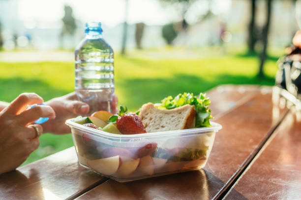 Woman's hands with a plastic container with sandwich, vegetables and fruits and a bottle of water on a table in a park in sunset