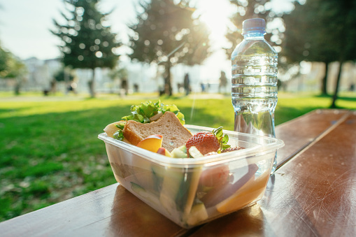 Plastic container with sandwich, vegetables and fruits and a bottle of water on a table in a park in sunset