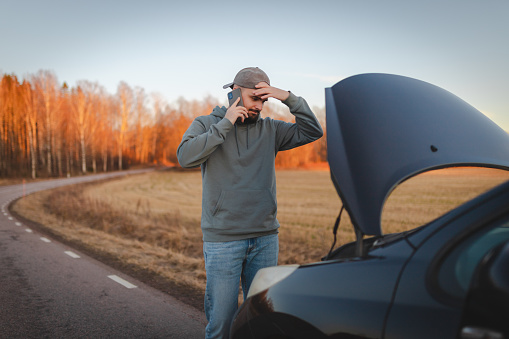 Stressed man opening the hood of his car on the road and calling for help.