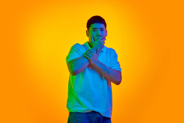 istock Surprised young Asian man with hands on mouth, standing in vibrant neon light against gradient orange background. 2075670159