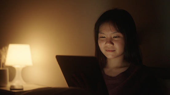 Teenage girl reading ebook on digital tablet in bedroom at night.