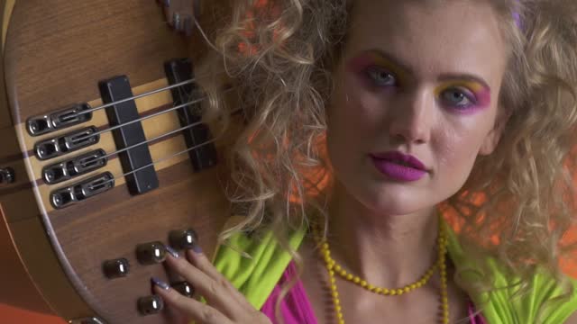 Blonde woman with bright retro style make up, shaking her head and strumming the guitar