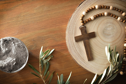 Christian cross on wooden base on a table with olive leaves and container full of ashes. Top view.
