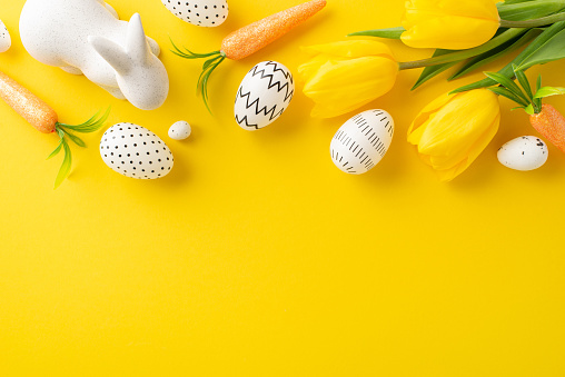 Easter inspiration idea. Overhead shot of simply colored eggs, cute bunny figure, carrots for the Easter Rabbit, and tulips on a bright yellow canvas with space for text or promotion