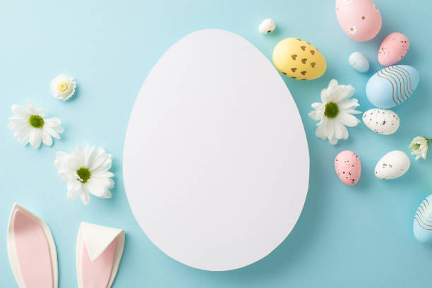 istock Easter art theme: top view of colorful eggs, Easter Bunny's ears, and young chamomile on a light blue background, featuring a vacant egg-shaped slot for writing or promotions 2075743524