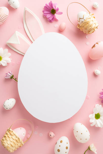 istock Trendy Easter idea capture. Overhead vertical view of eggs, tiny baskets, fresh chrysanthemums on a soft pink scene with rabbit ears sneaking behind vacant egg-shaped void for greeting words or advert 2075743540