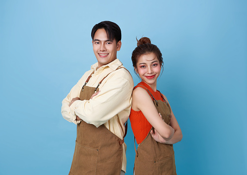 Portrait young asian barista couple partnership wearing apron standing and smile arms crossed back-to-back isolated on blue background.