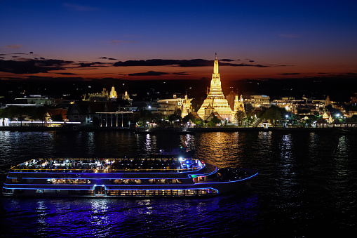 Stock photo showing a view across Bangkok's Chao Phraya River waterfront of Wat Arun Ratchawararam Ratchawaramahawihan (temple of dawn) illuminated at night.