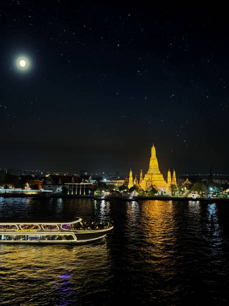 Image of dinner cruise boat passing Wat Arun (temple of dawn) viewed across Chao Phraya River, illuminated waterfront view, Bangkok, Thailand, reflections in water at night, moon and starlight sky background, focus on foreground