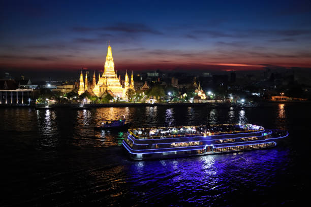 Stock photo showing a view across Bangkok's Chao Phraya River waterfront of Wat Arun Ratchawararam Ratchawaramahawihan (temple of dawn) illuminated at night.