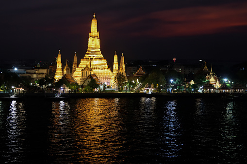 Stock photo showing a view across Bangkok's Chao Phraya River waterfront of Wat Arun Ratchawararam Ratchawaramahawihan (temple of dawn) illuminated at night.