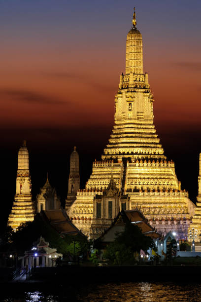 Stock photo showing a view across Bangkok's Chao Phraya River waterfront of Wat Arun Ratchawararam Ratchawaramahawihan (temple of dawn) illuminated at night.