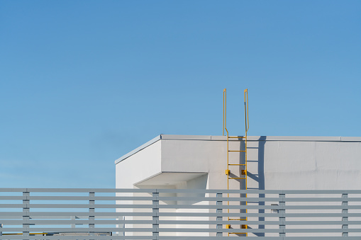 The photo presents a minimalist architectural scene, where the corner of a white building is accentuated by a striking yellow ladder, stark against the deep blue sky. The lines of the ladder and the details of the railing create a refined image beneath the tranquil expanse of blue, showcasing the allure of minimalist design.