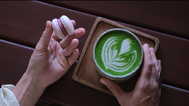 Macaron dessert and healthy matcha in hands in cozy coffee shop. Elegant hands carefully hold exquisite dessert and aromatic drink. Cinematic frame for coffee shops, pastry shops, blogs about desserts