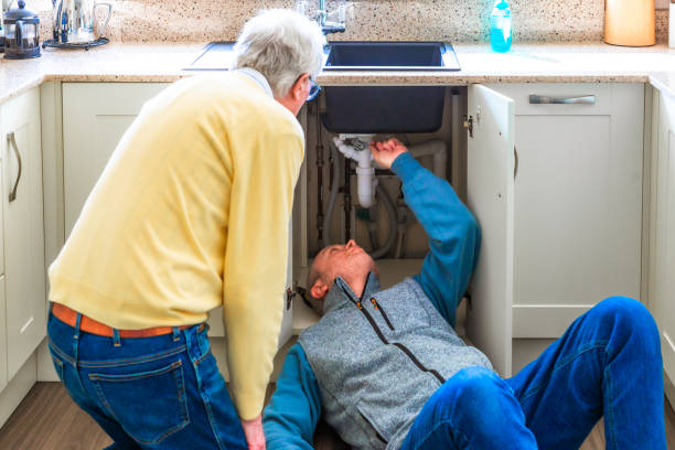 Plumber looking at pipes under kitchen sink with home owner observing