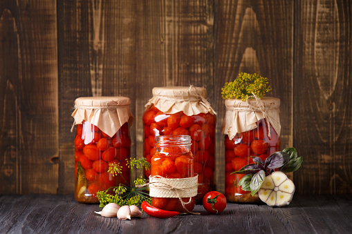 Canned cherry tomatoes in a closed and open jars, spices and herbs for marinade on a wooden background, copy space.