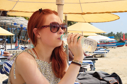 Stock photo showing close-up, profile view of red haired woman sitting on sun lounger under beach umbrella on sunny beach, drinking water from clear plastic bottle.