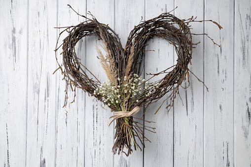 Crafted Wicker heart shaped wreath of birch branches hanging on white aged wooden background. Handmade DIY decoration