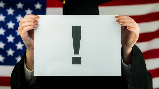 Graduate holds a poster with an exclamation point against the background of the American flag.
