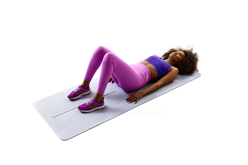 Angle view of young Africa-American woman lying on exercise mat and training abdominal against white studio background. Concept of sport, mourning routine, active and healthy lifestyle, energy, action