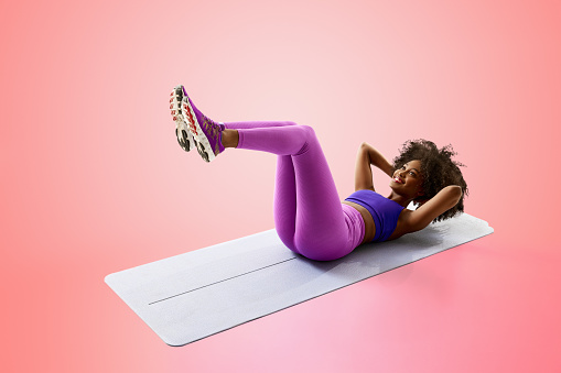 Young African-American woman doing abdominal exercises raising hands lying on exercise mat against gradient pink background. Concept of sport, mourning routine, active and healthy lifestyle, action.