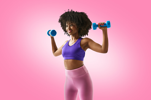 Smiling woman with dumbbells raised, wearing a purple sports bra and leggings against gradient pink studio background. Concept of sport, mourning routine, active and healthy lifestyle, energy, action.