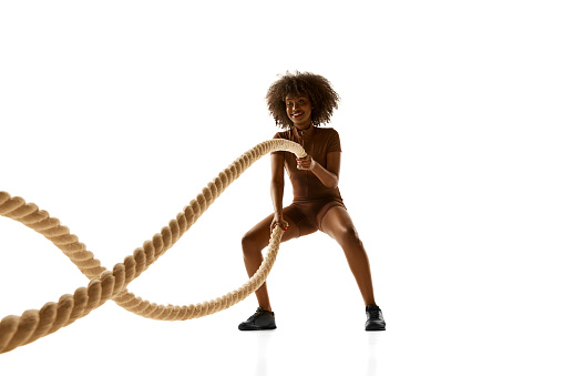 Smiling African-American woman in brown sports suit using thick ropes for fitness training against white studio background. Concept of sport, mourning routine, active and healthy lifestyle, energy.
