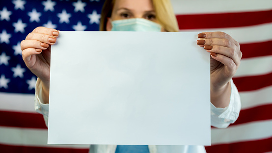 A masked woman holds an empty poster, stands against the background of the American flag.