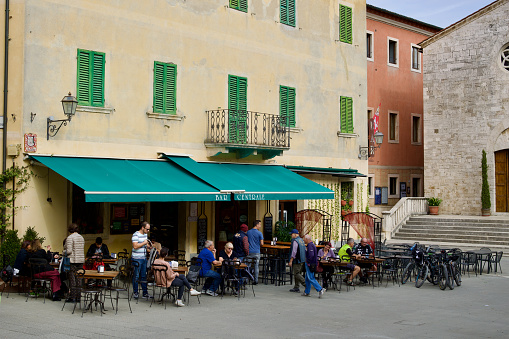 San Quirico d'Orcia, Province of Siena, Tuscany, Italy - 25 October 2022: Italian people sitting in a bar and enjoying life. Surrounding the square are more cafes, shops, and Chiesa di San Francesco. This church, also known as Chiesa della Madonna, is dedicated to Saint Francis and inside you can see a statue of the Madonna that was created by Andrea della Robbia.