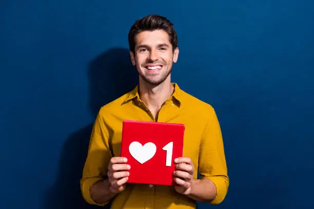 Photo of Photo portrait of attractive young man hold heart icon influencer wear trendy yellow clothes isolated on dark blue color background