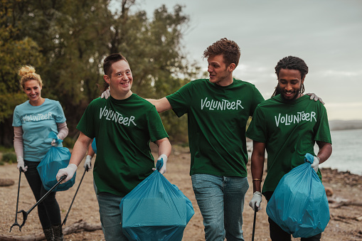 Three male volunteers in uniforms, one Caucasian, one African-American and one with down syndrome are walking by the beach, collecting trash and talking while their colleagues follow